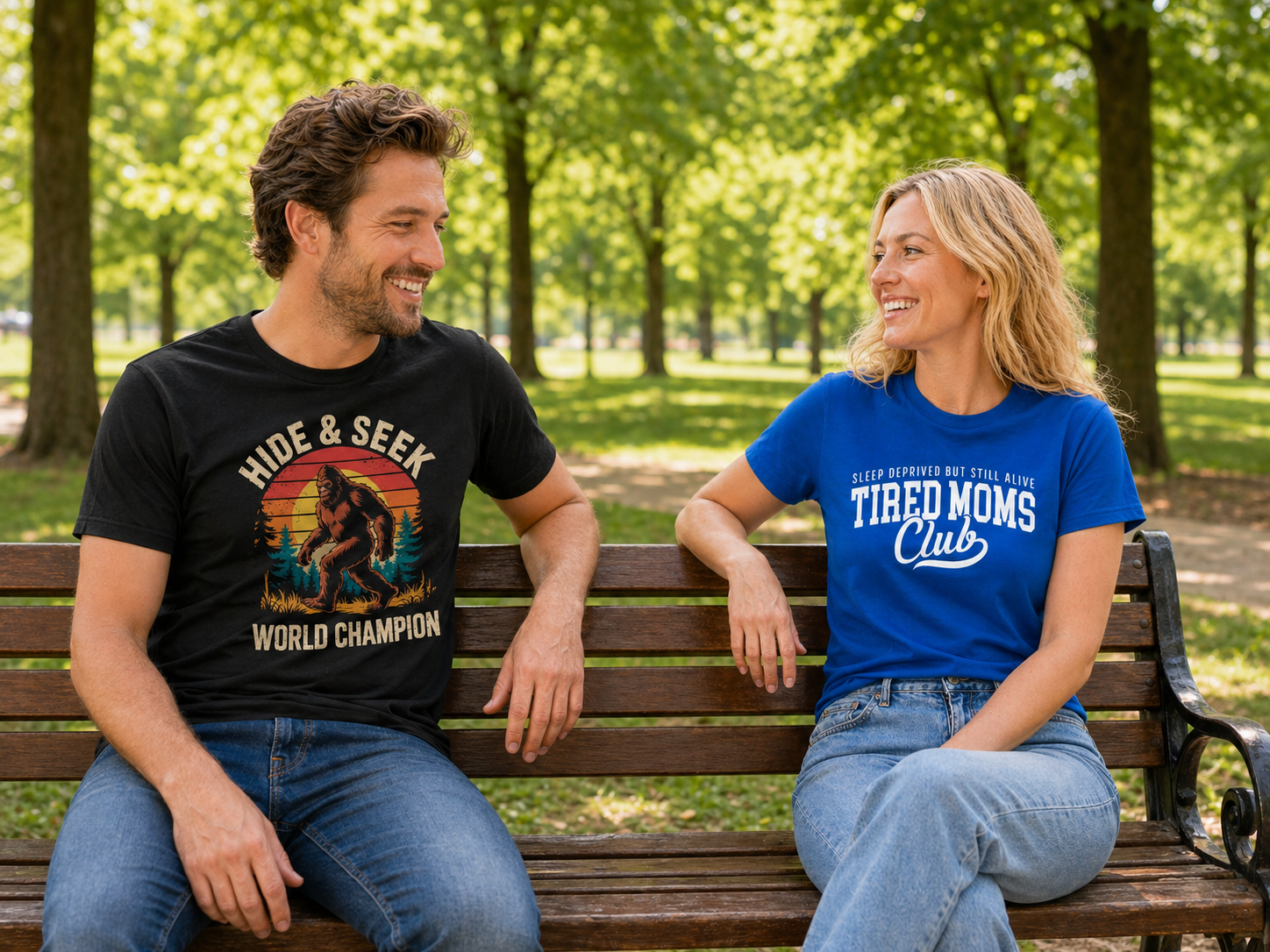 Happy couple sitting on a park bench modeling Laughing Designs funny t-shirts, including a black Bigfoot Hide and Seek World Champion tee and a blue Tired Moms Club shirt.