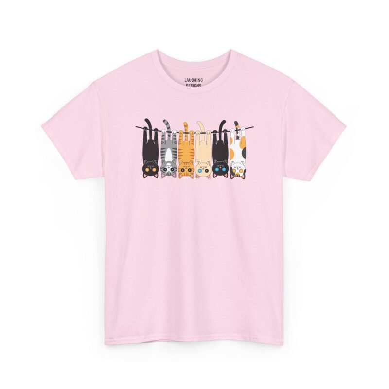 Hanging Cats on a Clothesline T-Shirt with six colorful cartoon cats dangling upside down from a black line, including a black cat with orange eyes, a grey tabby, an orange tabby, a cream cat, a black cat with blue eyes, and a calico cat, printed on a Light pink color cotton shirt.
