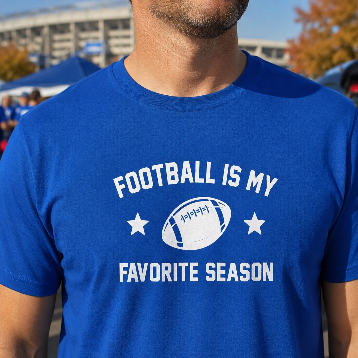 Football Is My Favorite Season T-Shirt by Laughing Designs, male model wearing a royal blue shirt with white text, star accents, and football graphic at a stadium tailgate.